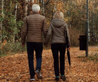 An older couple, seen from behind, walks hand in hand on a leaf-covered path, reflecting on Medicare benefits together.