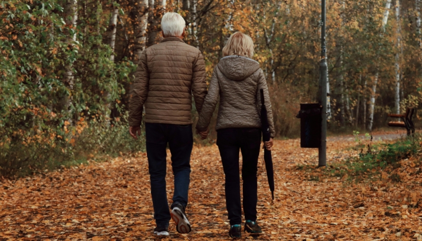 An older couple, seen from behind, walks hand in hand on a leaf-covered path, reflecting on Medicare benefits together.
